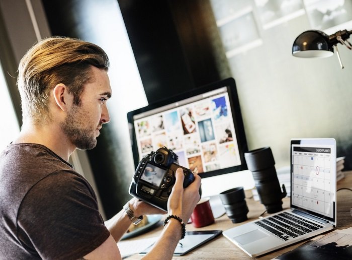 Hombre que sostiene la cámara frente a una computadora portátil y un monitor