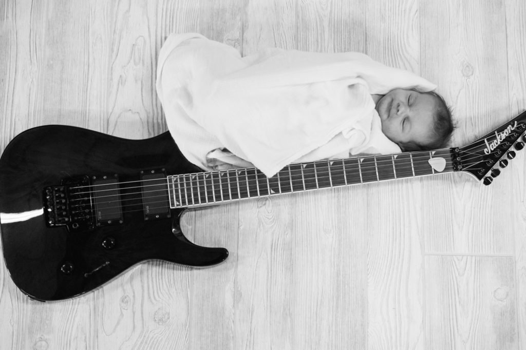 foto en blanco y negro de un bebé recién nacido envuelto en una manta blanca junto a una guitarra negra, tomada desde arriba. fotografía de recién nacido
