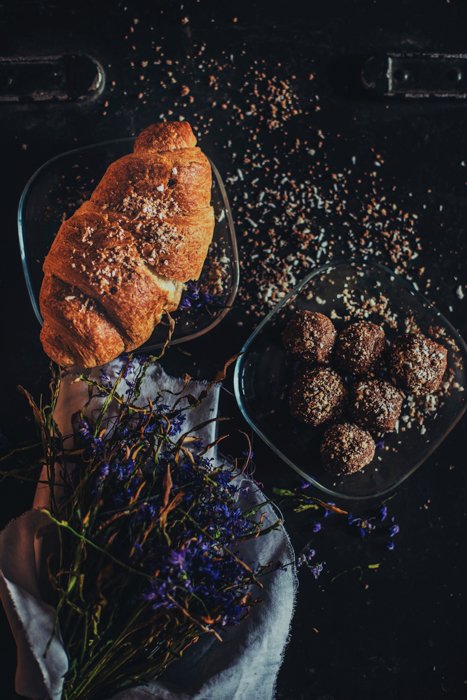 baguette y un plato de albóndigas sobre una mesa oscura con flores azules secas, migas esparcidas por todo