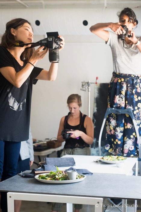 mujeres sosteniendo cámaras DSLR de pie en sillas, fotografiando comida en una mesa