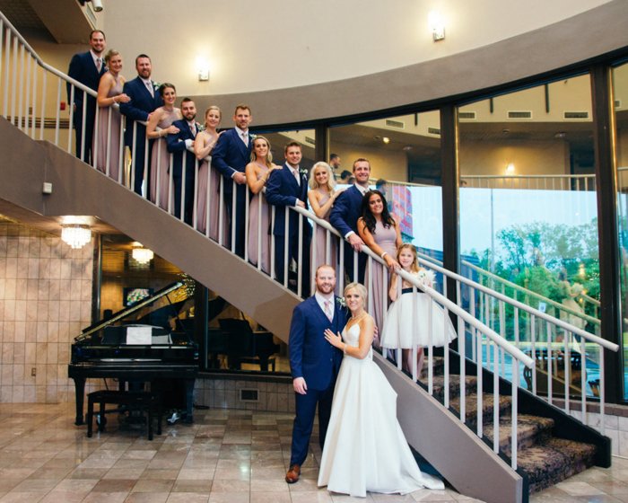 Foto de grupo grande de un séquito de bodas posando en una gran escalera curva, la novia y el novio en la parte inferior al frente, un piano de media cola cerca