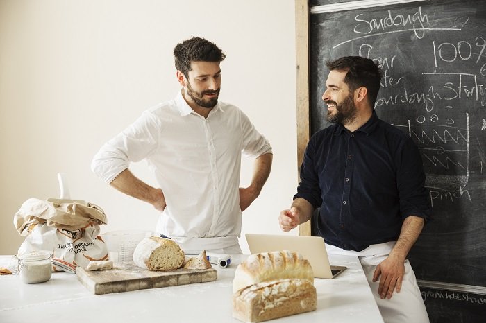 Dos panaderos parados en una mesa, usando una computadora portátil, pan recién horneado, una pizarra en la pared.