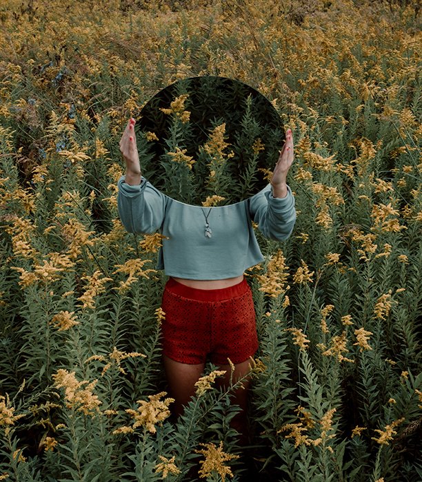 Fotografía de espejo surrealista de una modelo femenina en un campo cubierto de maleza, sosteniendo un espejo sobre su as que refleja más follaje cubierto de maleza