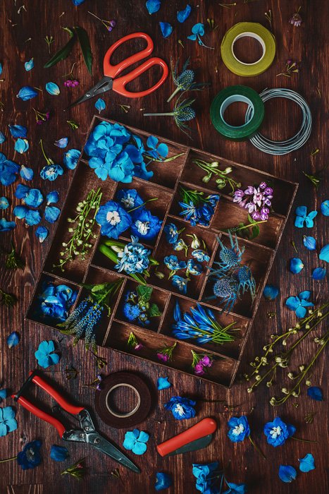 accesorios para alternativas creativas de fondo fotográfico: flores azul cielo, pétalos y hojas rosas y púrpuras en una caja de madera, más pétalos y hojas en un fondo de mesa de madera, con tijeras, cortaúñas y cinta adhesiva