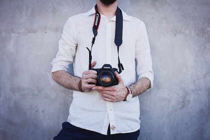 una imagen de un fotógrafo sosteniendo una cámara Canon DSLR de pie contra una pared gris