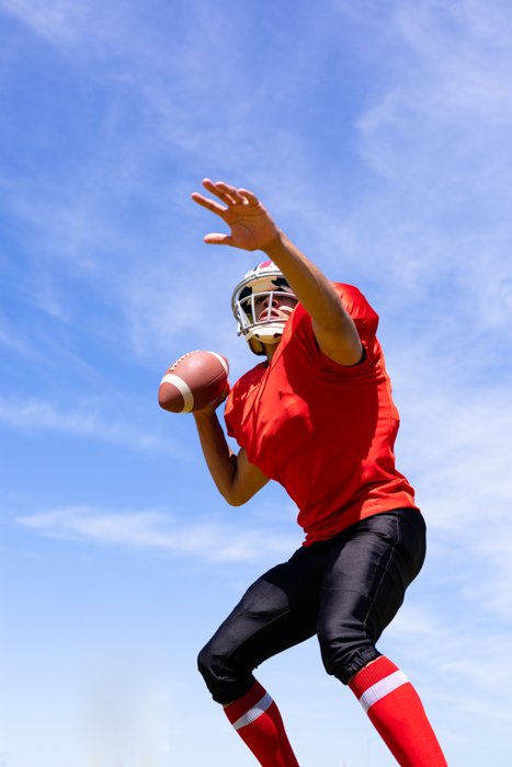 Vista frontal de ángulo bajo de un jugador de fútbol americano masculino de raza mixta con uniforme de equipo, entrenando en un campo deportivo, preparándose para lanzar una pelota de fútbol, ​​con el cielo azul en el fondo