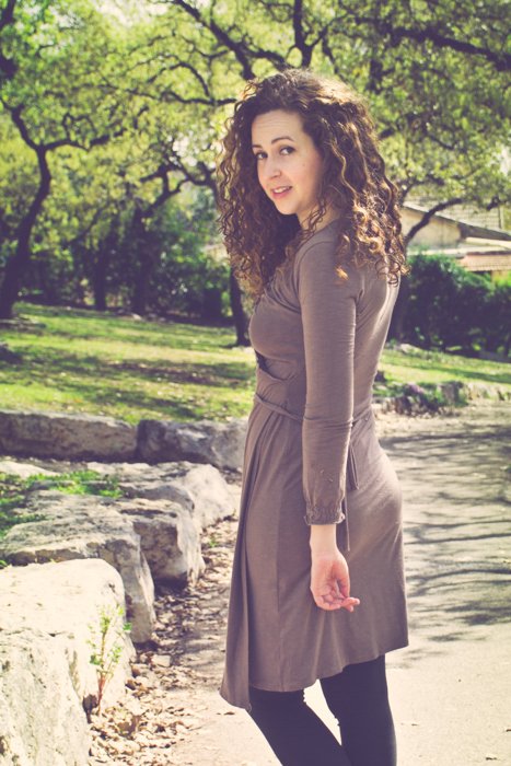 foto recortada de una morena de cabello rizado con un vestido marrón y botas mirando hacia atrás mientras camina por un camino de tierra en un parque