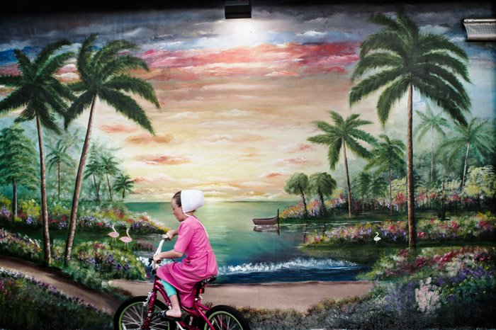 Foto de una niña en bicicleta frente a un paisaje pintado por Dina Litovsky.  Fotógrafos famosos a seguir.