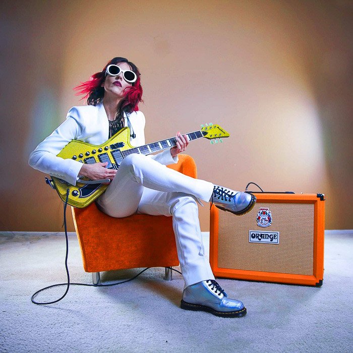 Una mujer sentada en una silla naranja junto a un amplificador naranja sosteniendo una guitarra eléctrica