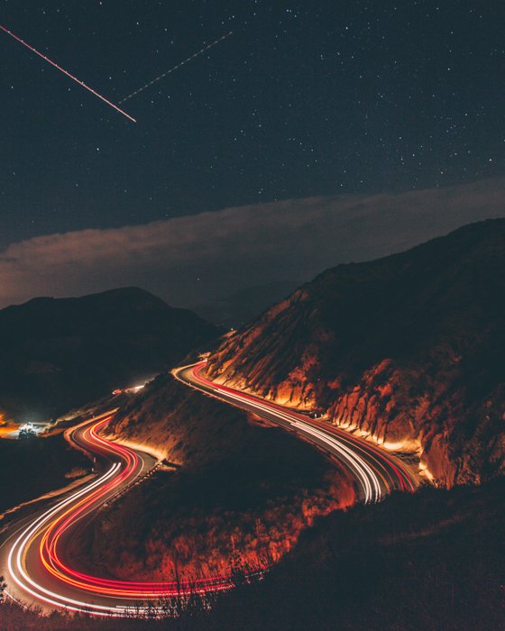 una foto de lapso de tiempo de autos conduciendo en una carretera de montaña curva con estrellas fugaces en el cielo